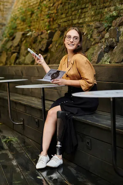 En un encantador entorno de cafetería, una joven con una pierna protésica se involucra con su teléfono, irradiando alegría y confianza. - foto de stock