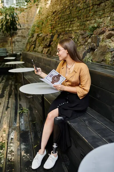 A young woman with a prosthetic leg sits relaxed in a cafe, savoring her drink and scrolling on her phone, embracing life. — Stock Photo