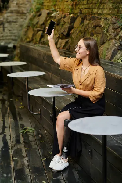 A young woman with a prosthetic leg smiles brightly while using her phone in a charming cafe setting. — Stock Photo
