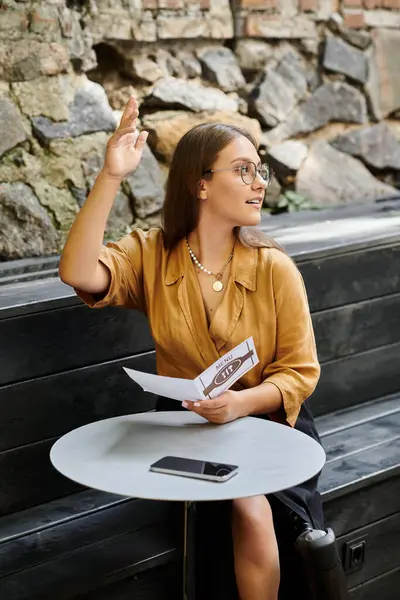 A young woman in a stylish top sits at a cafe table, smiling and asking for assistance, embracing her daily routine. — Stock Photo