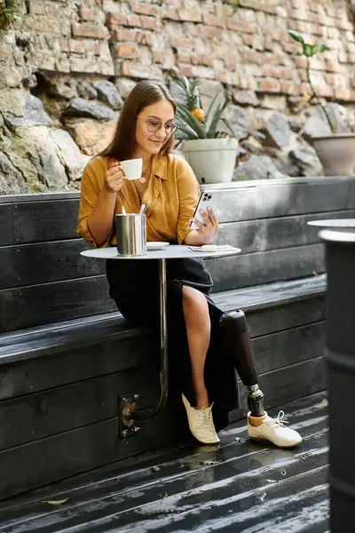 In un caffè alla moda, una giovane donna con una gamba protesica sorseggia caffè mentre si impegna con il suo telefono, abbracciando la vita. — Foto stock