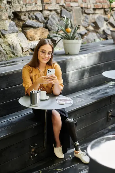 A young woman with a prosthetic leg sits comfortably in a cafe, engaged with her phone and savoring a warm drink. — Stock Photo