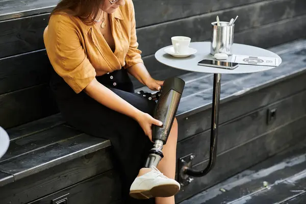 A young woman sits comfortably in a cafe, sipping coffee and adjusting her prosthetic leg, embracing her daily routine. — Stock Photo
