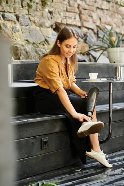 A young woman with a prosthetic leg sits in a cafe, sipping coffee while comfortably adjusting her leg, embodying a relaxed lifestyle. — Stock Photo