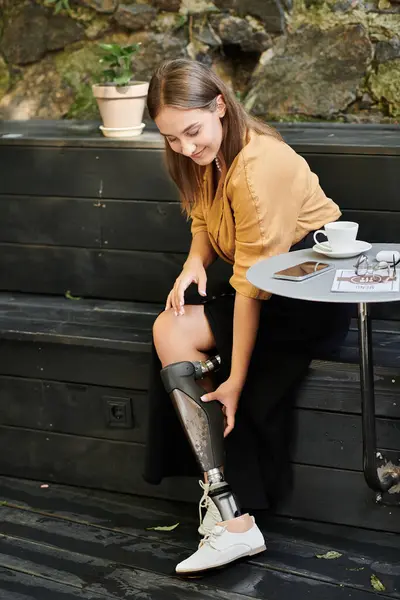 In a delightful cafe, a young woman smiles while adjusting her prosthetic leg, embracing everyday life with style. — Stock Photo