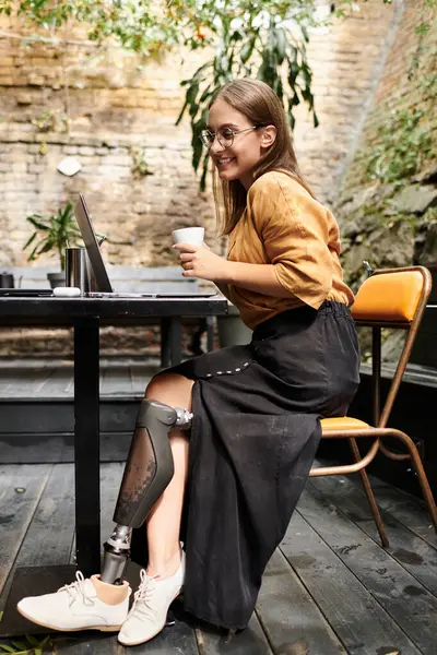 In un affascinante caffè, una giovane donna con un arto artificiale assapora il caffè e si impegna con il suo computer portatile, abbracciando la vita quotidiana.. — Stock Photo