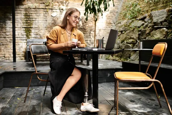 A young woman with an artificial limb sits comfortably in a cafe, sipping coffee while working on her laptop, embracing her daily routine. — Stock Photo