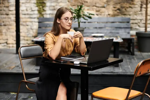 Una giovane donna si rilassa in un caffè, sorseggiando il suo drink e lavorando sul suo computer portatile, abbracciando la sua routine quotidiana con stile. — Foto stock