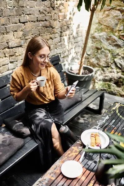 Una joven se sienta cómodamente en un café, tomando café y revisando su teléfono mientras disfruta de un dulce. - foto de stock