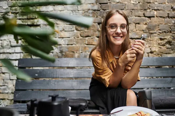A young woman sits at a cafe, smiling while holding a smartphone, showcasing her vibrant daily life and confidence. — Stock Photo