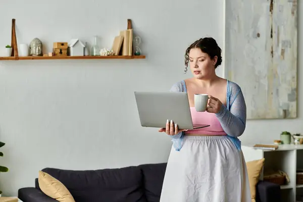 A plus size woman interacts with her laptop, holding a mug, in a comfortable home setting. — Stock Photo