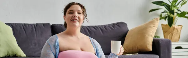Веселая женщина plus size в уютной домашней одежде, держа чашку чая в руках, дома. — стоковое фото