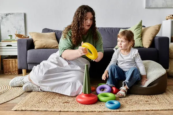 Eine Plus-Size-Mutter verwickelt ihre Tochter in ein lustiges, buntes Stapelspiel, während sie gemütlich sitzt. — Stock Photo