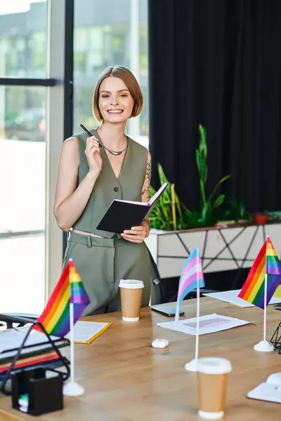 Uma mulher sorri orgulhosamente enquanto segura um caderno, cercado por bandeiras de orgulho e xícaras de café. — Fotografia de Stock