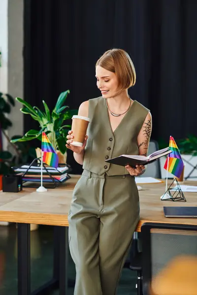 A woman enjoys coffee while reviewing notes in an inclusive office setting with pride flags. — Stock Photo