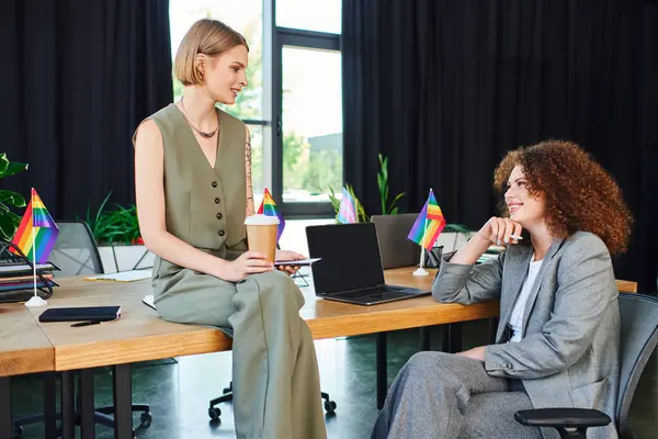 Two coworkers laugh and engage at their desks, surrounded by colorful flags symbolizing pride. — Stock Photo