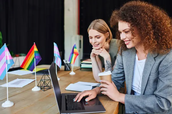 In a lively office, two colleagues joyfully share a laptop, displaying pride flags. — Stock Photo
