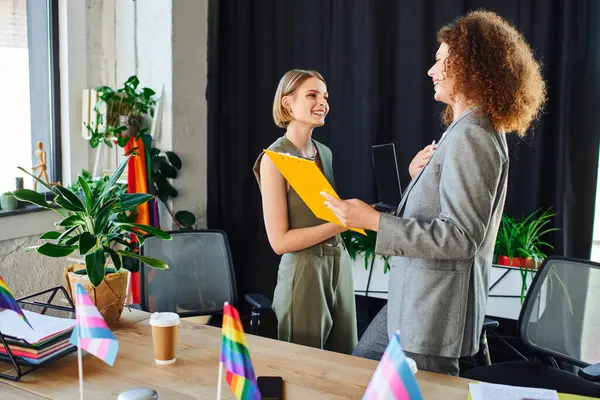 Two coworkers share a joyful moment in their inclusive office environment surrounded by flags. — Stock Photo