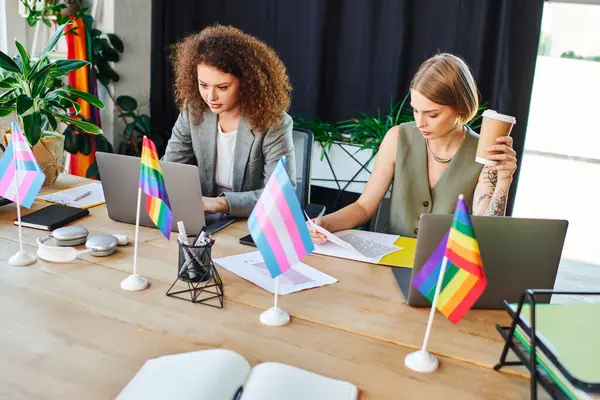 Two coworkers engage in a creative workspace adorned with vibrant flags celebrating inclusivity. — Stock Photo