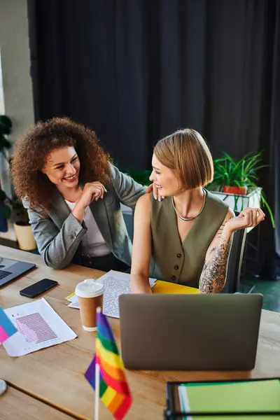 Dos compañeros de trabajo chatean animadamente en medio de vibrantes banderas de la comunidad LGBTQ. - foto de stock