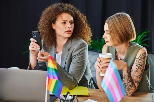 Zwei Mitarbeiter unterhalten sich nachdenklich, während sie von Stolz-Fahnen und Büromaterialien umgeben sind. — Stockfoto