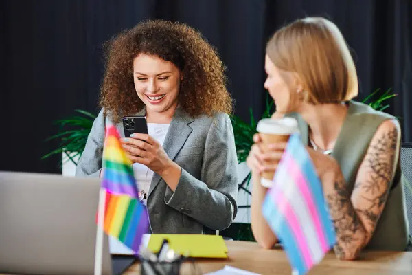 Dos colegas comparten un momento de luz, interactuando con sus teléfonos mientras muestran banderas de orgullo. - foto de stock