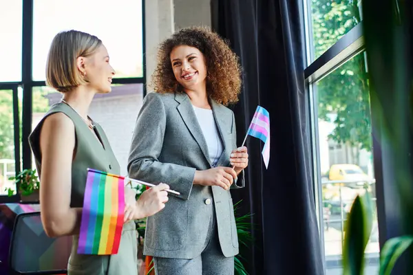 Two coworkers joyfully engage, waving rainbow flags in a bright, welcoming workspace. — Stock Photo