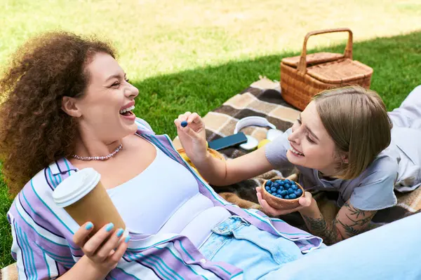 Two young women share laughter while relaxing on a picnic blanket with snacks in a sunny setting. — Stock Photo