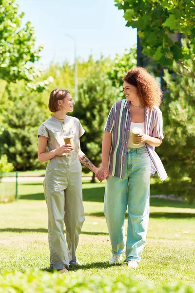 Two friends happily stroll together in a green park, sharing drinks and laughter under the sun. — Stock Photo