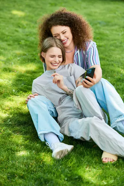 Two friends share a joyful moment on the grass, laughing and looking at a phone together. — Stock Photo