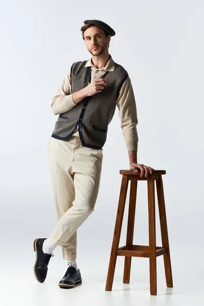 Un gentleman sophistiqué portant une tenue tendance se penche contre un tabouret rustique dans un studio. — Stock Photo