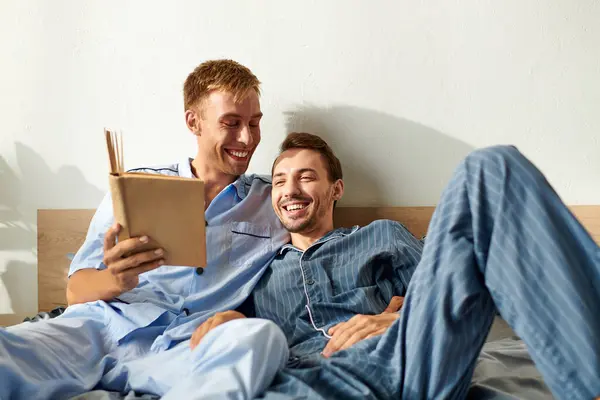 A joyful gay couple shares a warm moment, reading and laughing together in their cozy pajamas. — Stock Photo