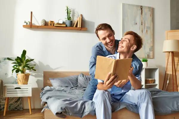 A joyful gay couple cuddles in their pajamas, sharing laughter and stories over a book in the morning light. — Stock Photo