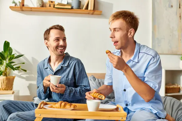 A loving gay couple in pajamas share breakfast and laughter, embracing a peaceful morning at home. — Stock Photo
