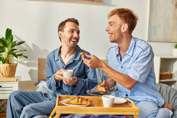 A loving gay couple enjoys a cozy morning together, sipping coffee and sharing pastries in their pajamas. — Stock Photo