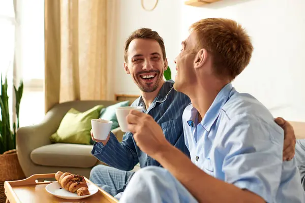 A joyful gay couple enjoys a warm morning together, sharing smiles and drinks in their comfy pajamas. — Stock Photo