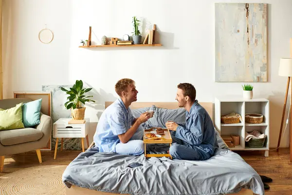 A joyful gay couple enjoys a leisurely morning together in pajamas, sharing breakfast and laughter in bed. — Stock Photo