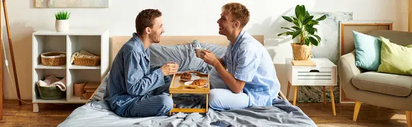 A loving gay couple enjoys breakfast together in their pajamas, sharing smiles and affection in the morning light. — Stock Photo