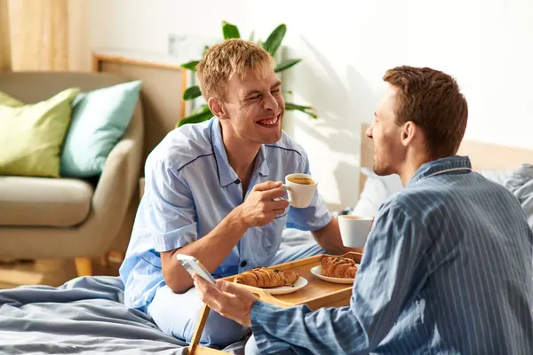 Two men in pajamas enjoying breakfast together, sharing laughter and affection in their bright bedroom. — Stock Photo