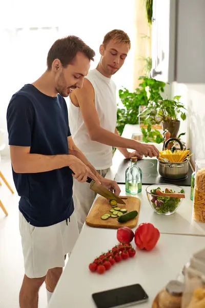 In a bright kitchen, two men happily prepare a fresh salad, showcasing love through cooking together. — Stock Photo
