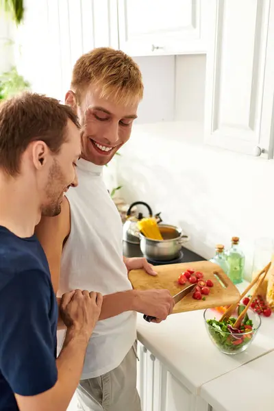 Two men are happily preparing a salad in their chic kitchen, slicing vegetables and sharing laughs. — Stock Photo