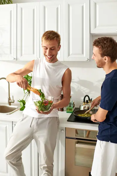 Due uomini assaporano un momento gioioso nella loro cucina elegante, facendo un'insalata fresca e godendosi reciprocamente la compagnia.. — Foto stock