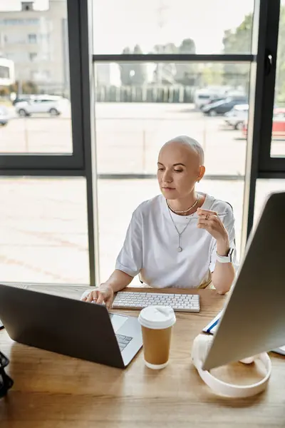 Una giovane donna calva in abiti eleganti si concentra sul suo computer portatile, sorseggiando caffè pensieroso. — Foto stock