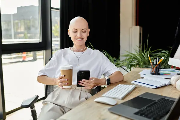 A young bald woman dressed elegantly sits at a desk, sipping coffee and browsing her phone. — Stock Photo