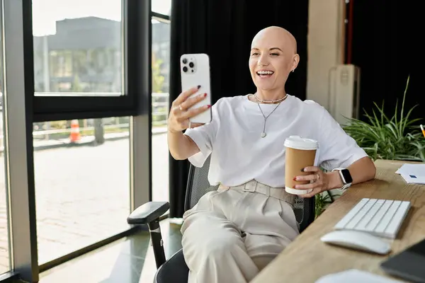 A young bald woman smiles brightly, holding coffee and video chatting in a stylish workspace. — Stock Photo