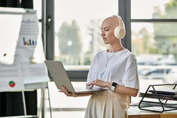 A young woman dressed elegantly interacts with her laptop, wearing headphones in a bright workspace. — Stock Photo