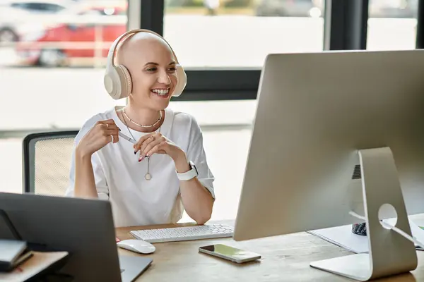 A young bald woman in stylish attire smiles while immersed in her workspace, wearing headphones. — Stock Photo