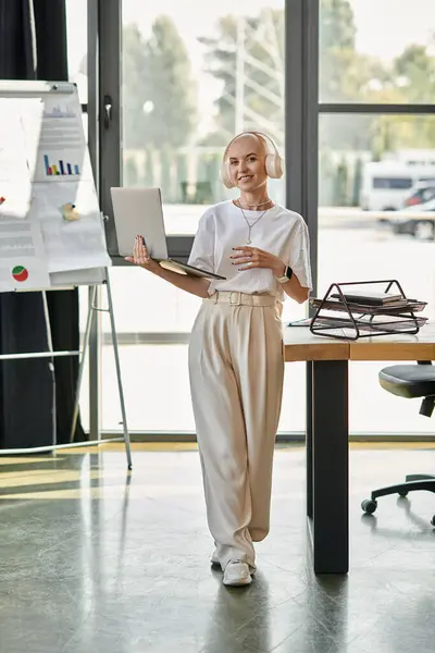 Una mujer calva joven con estilo se pone de pie con confianza en un traje elegante, la participación con un ordenador portátil. - foto de stock