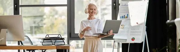 A young bald woman confidently presents her ideas while holding a laptop in a bright office. — Stock Photo