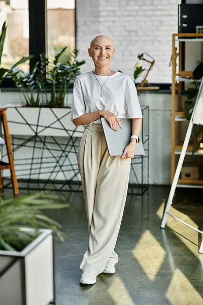 A young bald woman dressed elegantly stands in a stylish workspace, holding a laptop. — Stock Photo